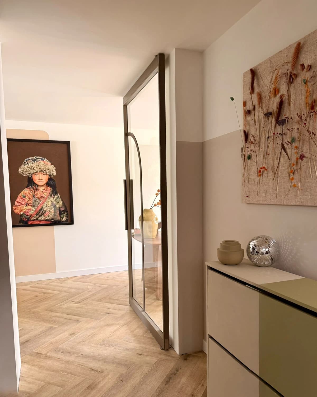 Stalen taatsdeur met glas en een boog in het deurblad, halfopen aan het eind van een gang met een visgraatvloer, met erachter een ingelijst portret